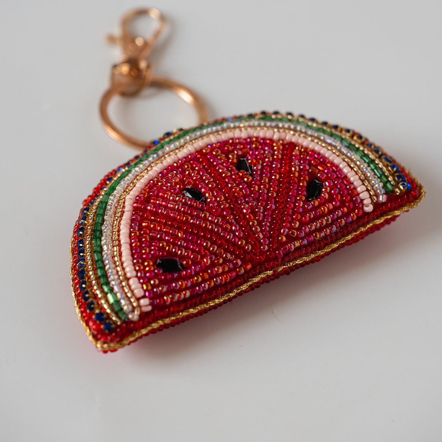A beaded watermelon slice keychain charm with a gold-tone clasp, featuring red, green, white, and black seed details on a light gray background.