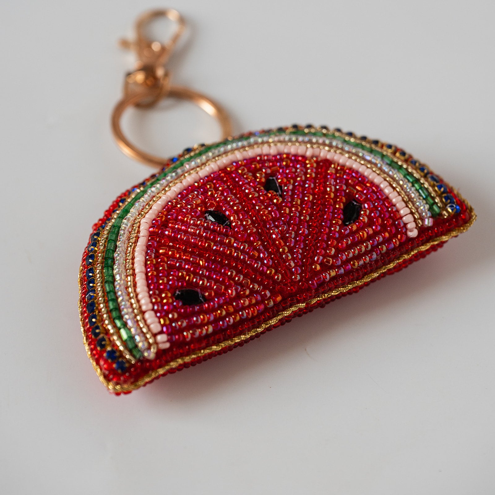 A beaded watermelon slice keychain charm with a gold-tone clasp, featuring red, green, white, and black seed details on a light gray background.