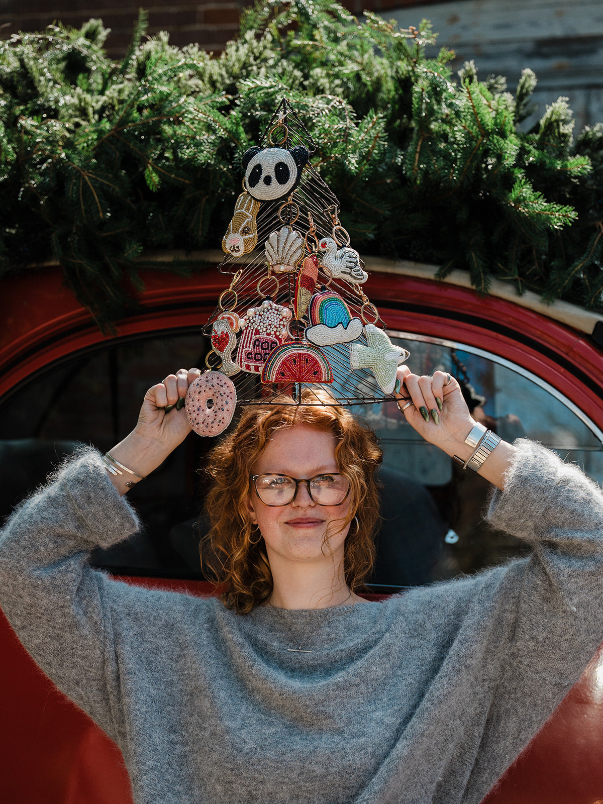 A woman with curly red hair and glasses holds up a decorative wire Christmas tree ornament featuring various cute illustrations like a panda, donut, and ice cream cone, set against a red vintage car window with green pine branches.