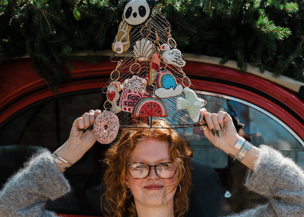 A woman with curly red hair and glasses holds up a decorative wire Christmas tree ornament featuring various cute illustrations like a panda, donut, and ice cream cone, set against a red vintage car window with green pine branches.