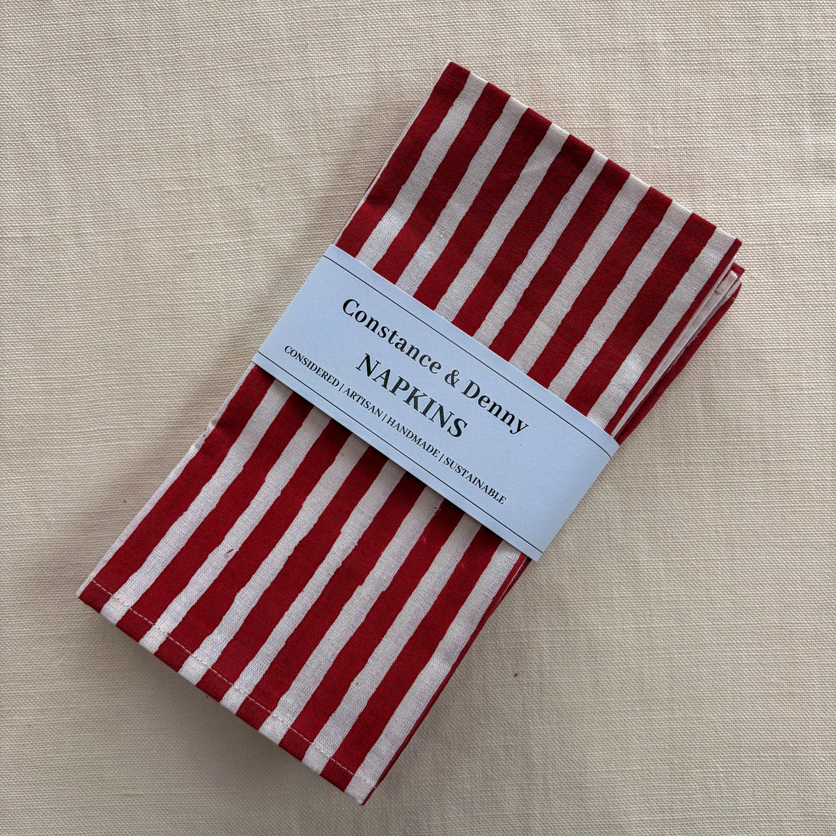A folded stack of red and white striped paper napkins with a white paper label reading 'Constance & Denny NAPKINS' on a beige linen background.