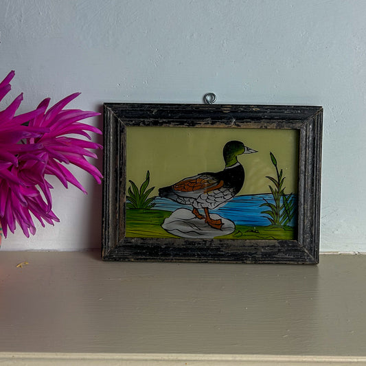 Framed artwork of a duck by a pond with a pink flower beside it on a white surface.