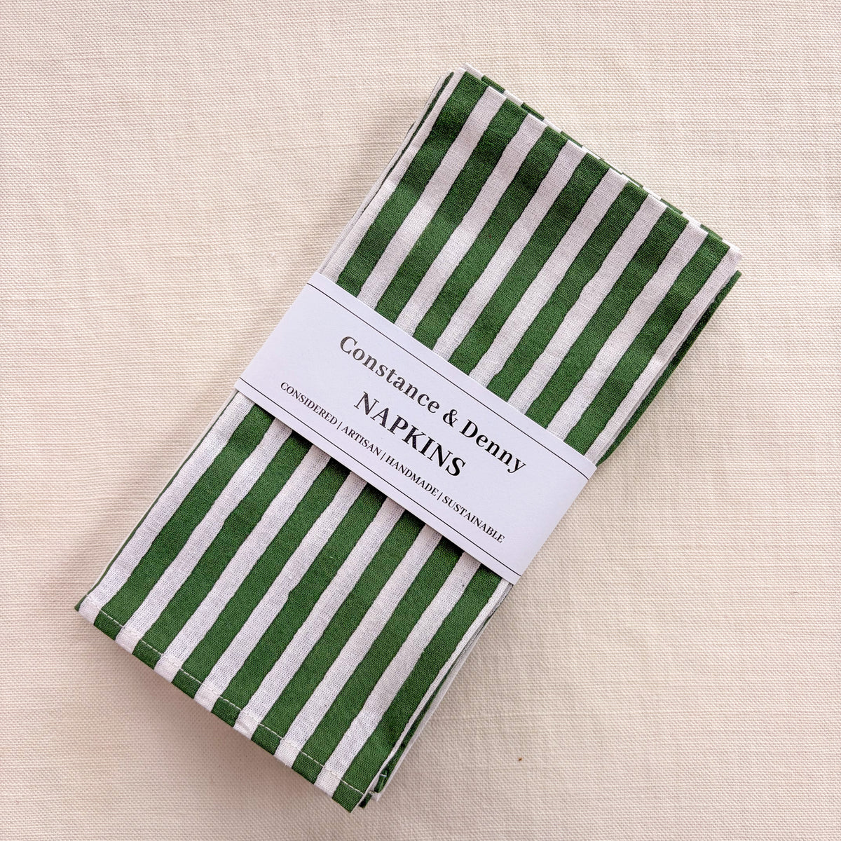 A folded set of Constance & Denny napkins with green and white vertical stripes, wrapped in a white paper band with the brand name printed on it, set against a neutral beige linen background.