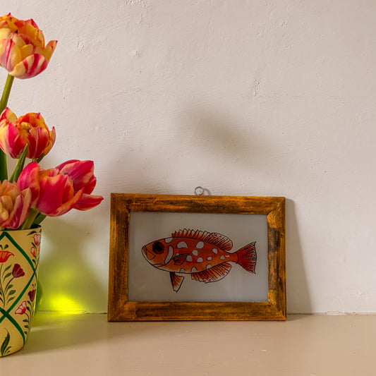 Framed illustration of a fish next to a vase with flowers on a white surface.