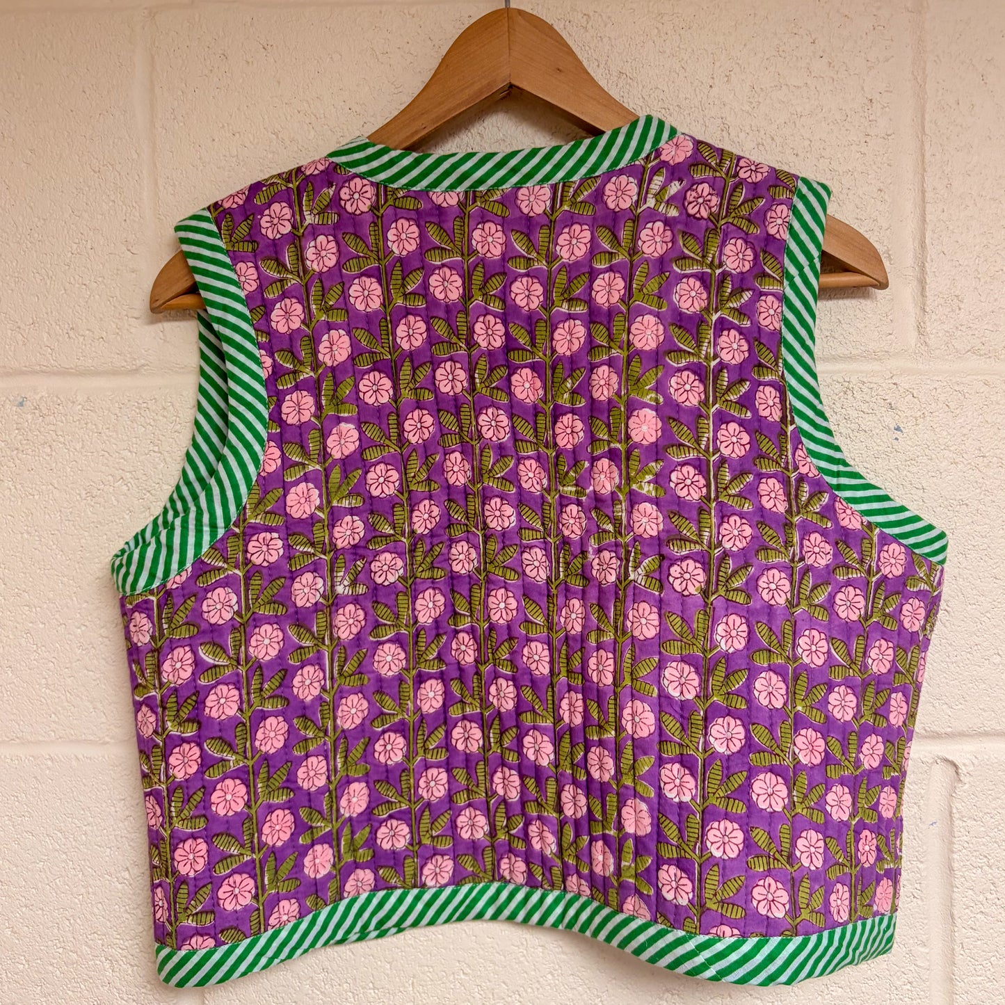 Purple floral-patterned waistocat with green and white striped edges on a wooden hanger against a beige wall.