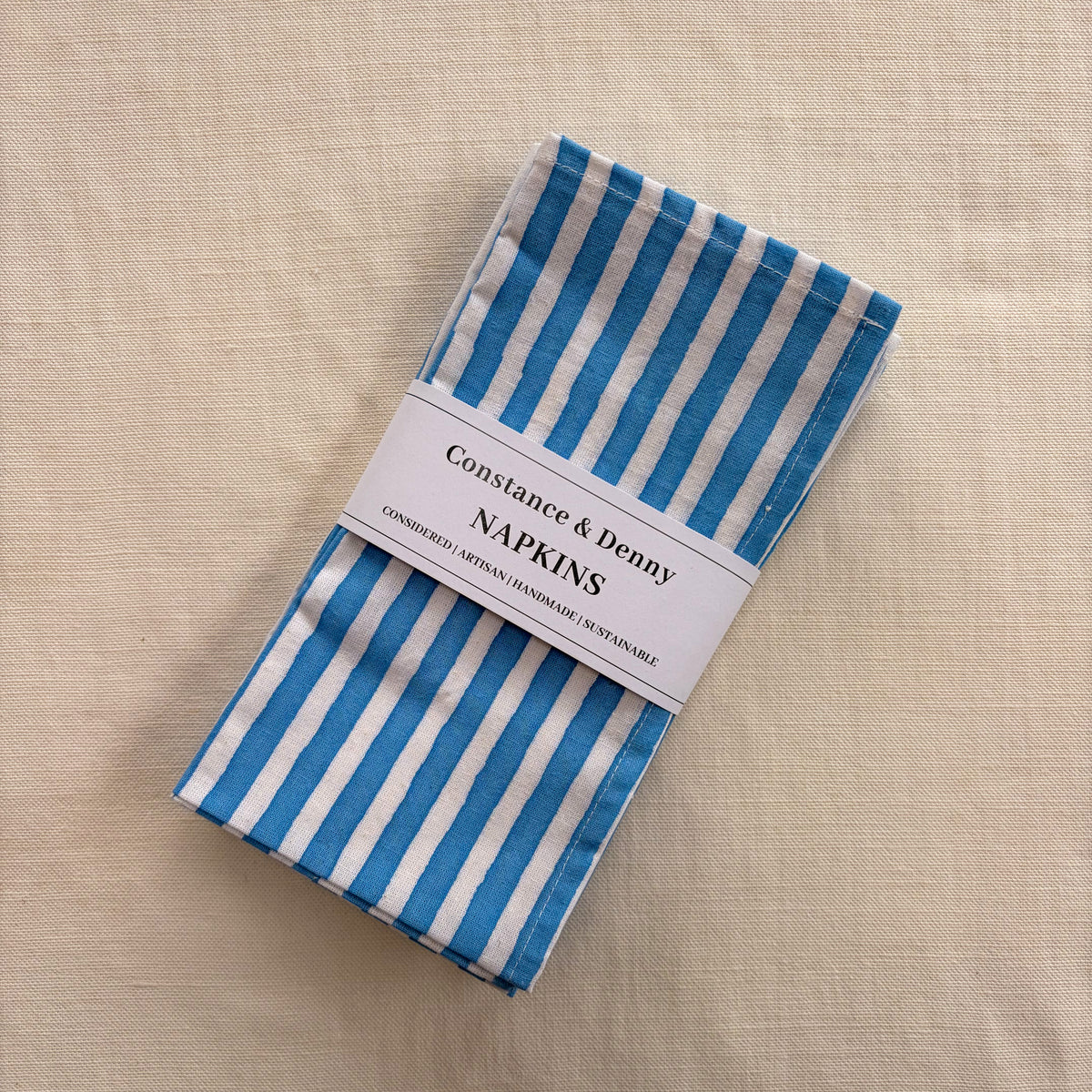 Blue and white striped paper napkins wrapped in a white paper band that reads 'Constance & Denny NAPKINS', placed on a textured beige linen surface.