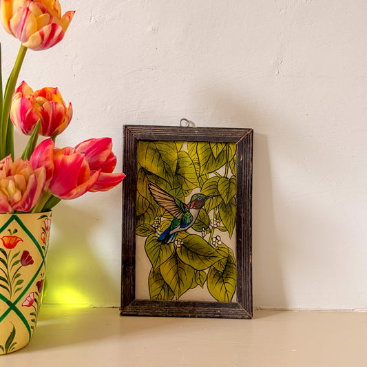 Framed artwork of a hummingbird and leaves next to a vase of tulips on a white surface.