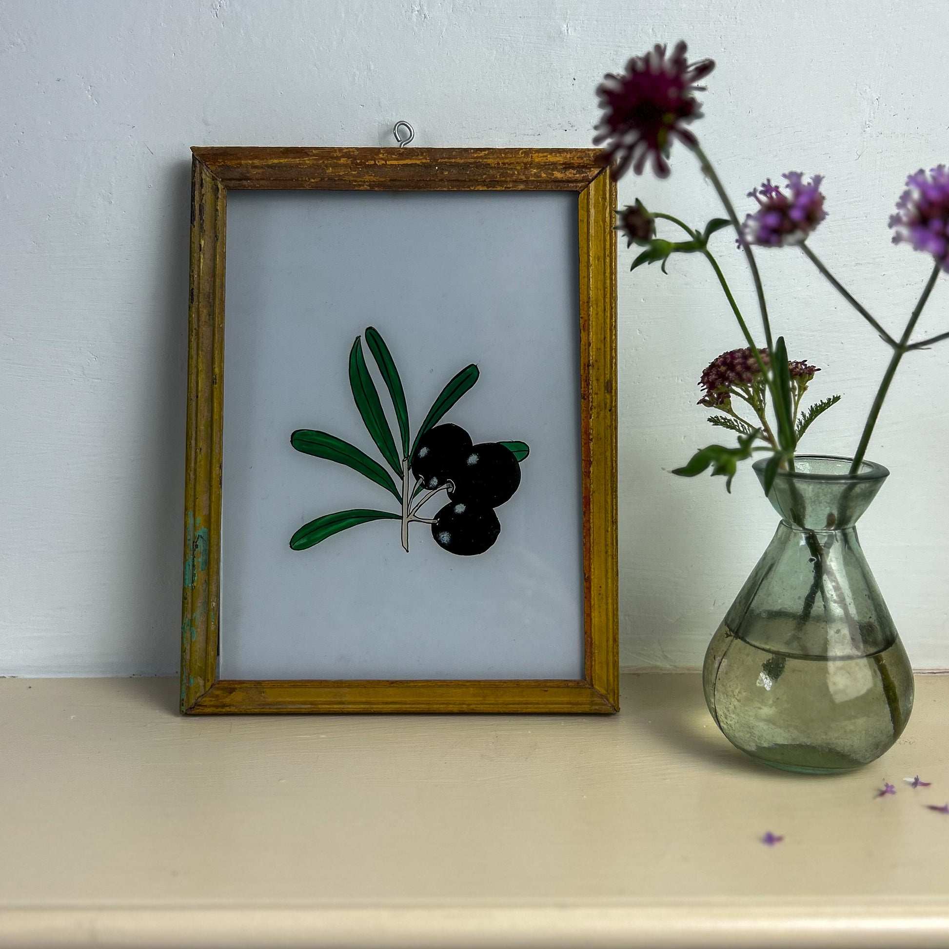 Framed artwork of olives next to a vase with flowers on a shelf.  indian-glass-painting-reverese-black-olive-branch