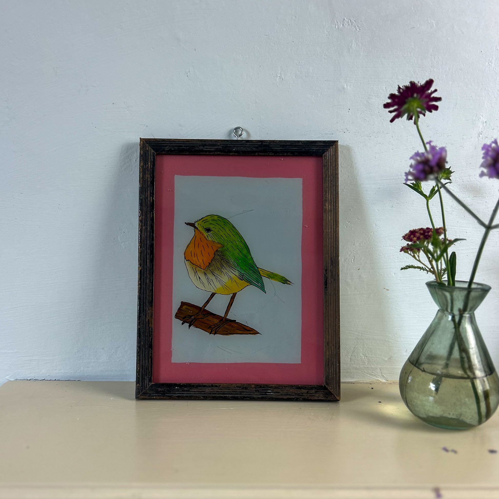 Framed artwork of a colorful bird on a table with a vase of flowers. indian-glass-painting-reverese-robin
