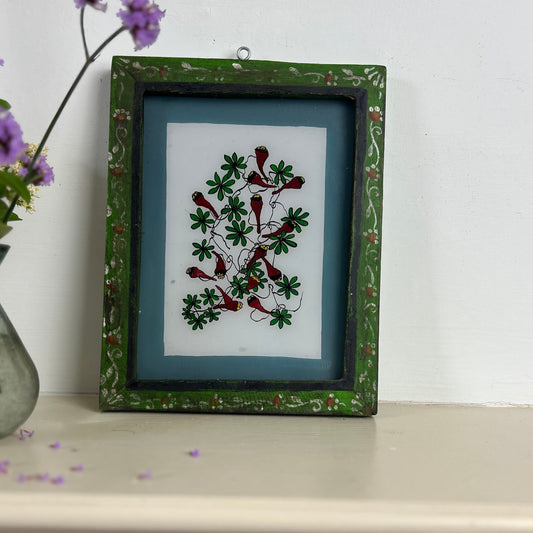 Framed artwork with floral design on a shelf with a vase of flowers. indian-glass-painting-reverse-chilli-plant