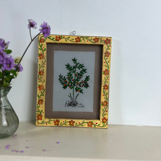 Decorative framed artwork with floral design on a shelf next to a vase with purple flowers. indian-glass-painting-reverse-orange-tree