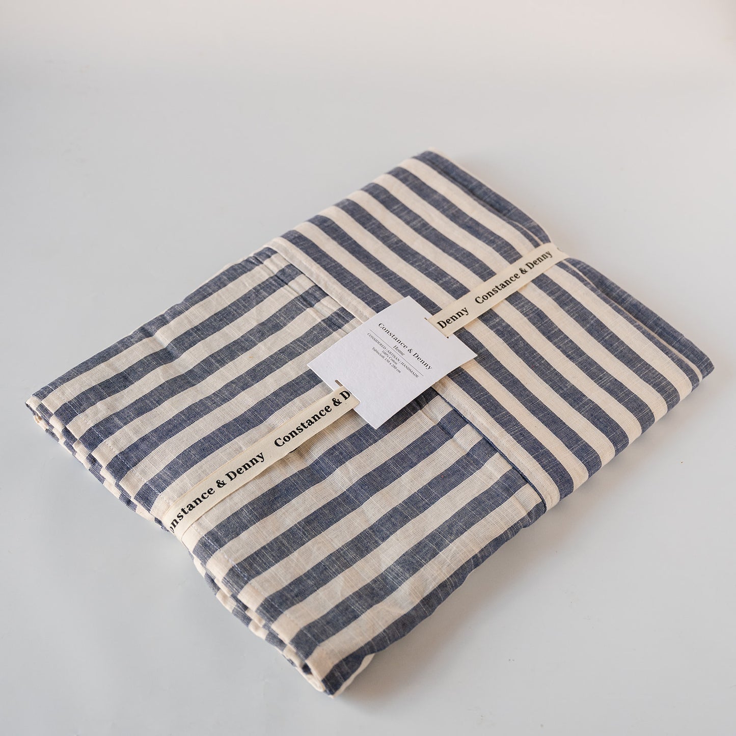 A folded striped textile, likely a blanket or throw, in navy blue and cream cotton fabric, wrapped with a paper band and tag. The brand 'Constance & Benny' is clearly printed on the paper band.