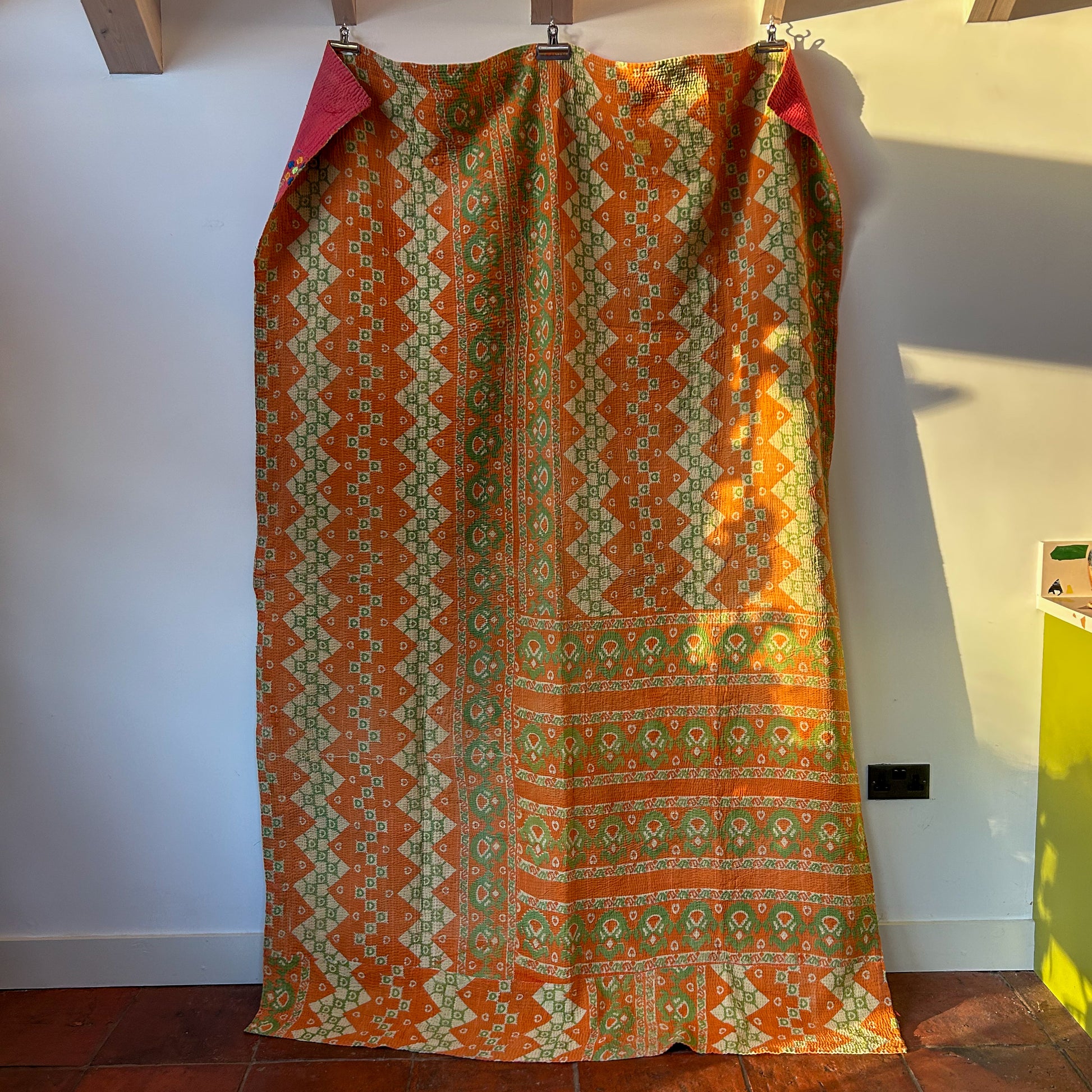 Traditional patterned kantha draped over a chair against a white wall.