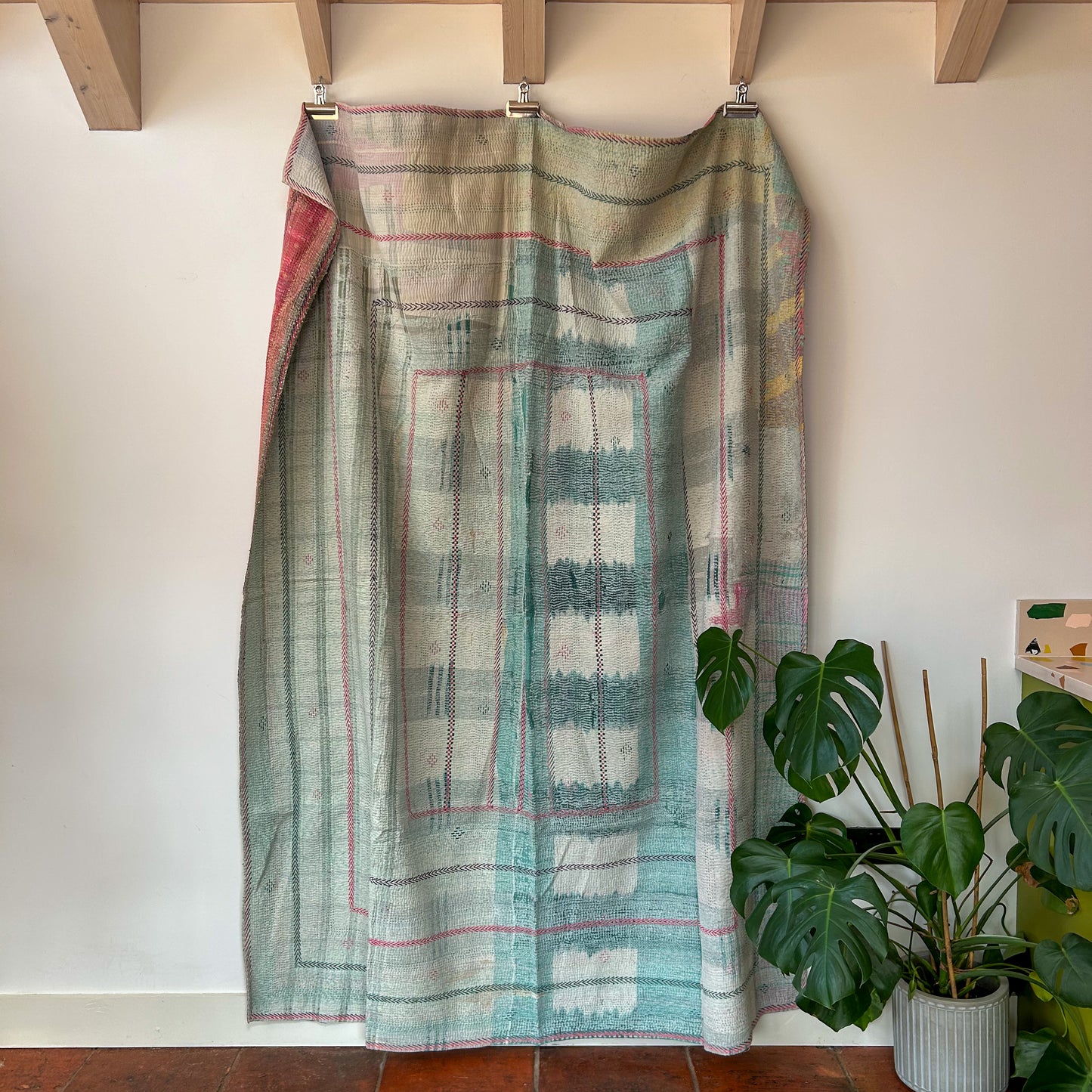 Textile with geometric pattern draped over a wooden frame against a white wall with a plant in the foreground. kantha-vintage-2027- hand-made-blanket-throw-textiles-wall-hanging