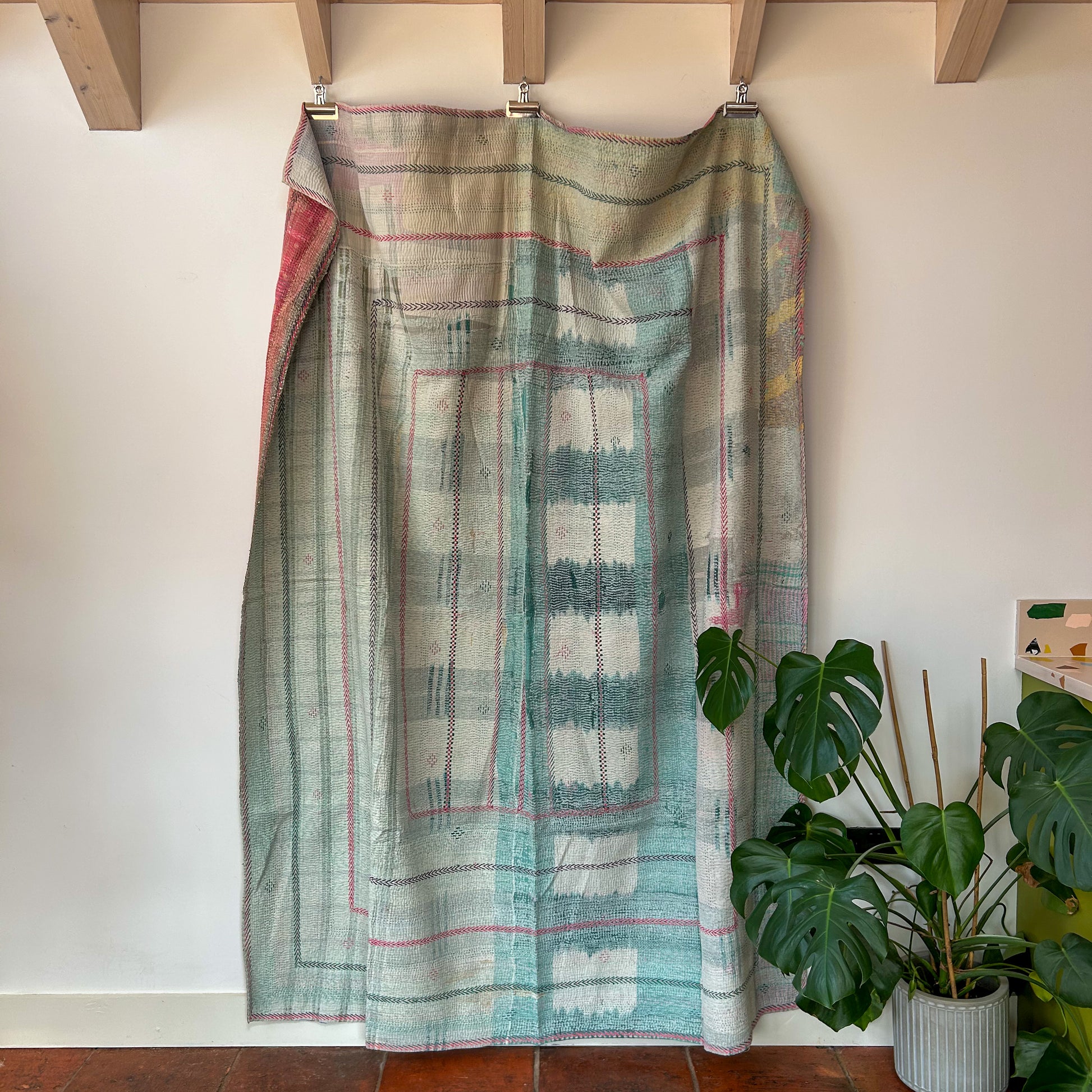 Textile with geometric pattern draped over a wooden frame against a white wall with a plant in the foreground. kantha-vintage-2027- hand-made-blanket-throw-textiles-wall-hanging