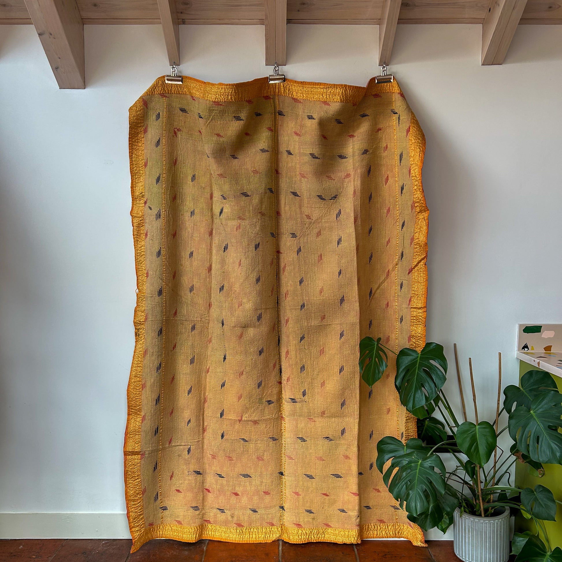 Yellow patterned curtain hanging against a white wall with a plant in the corner. kantha-vintage-2025-hand-made-blanket-throw-textiles-wall-hanging