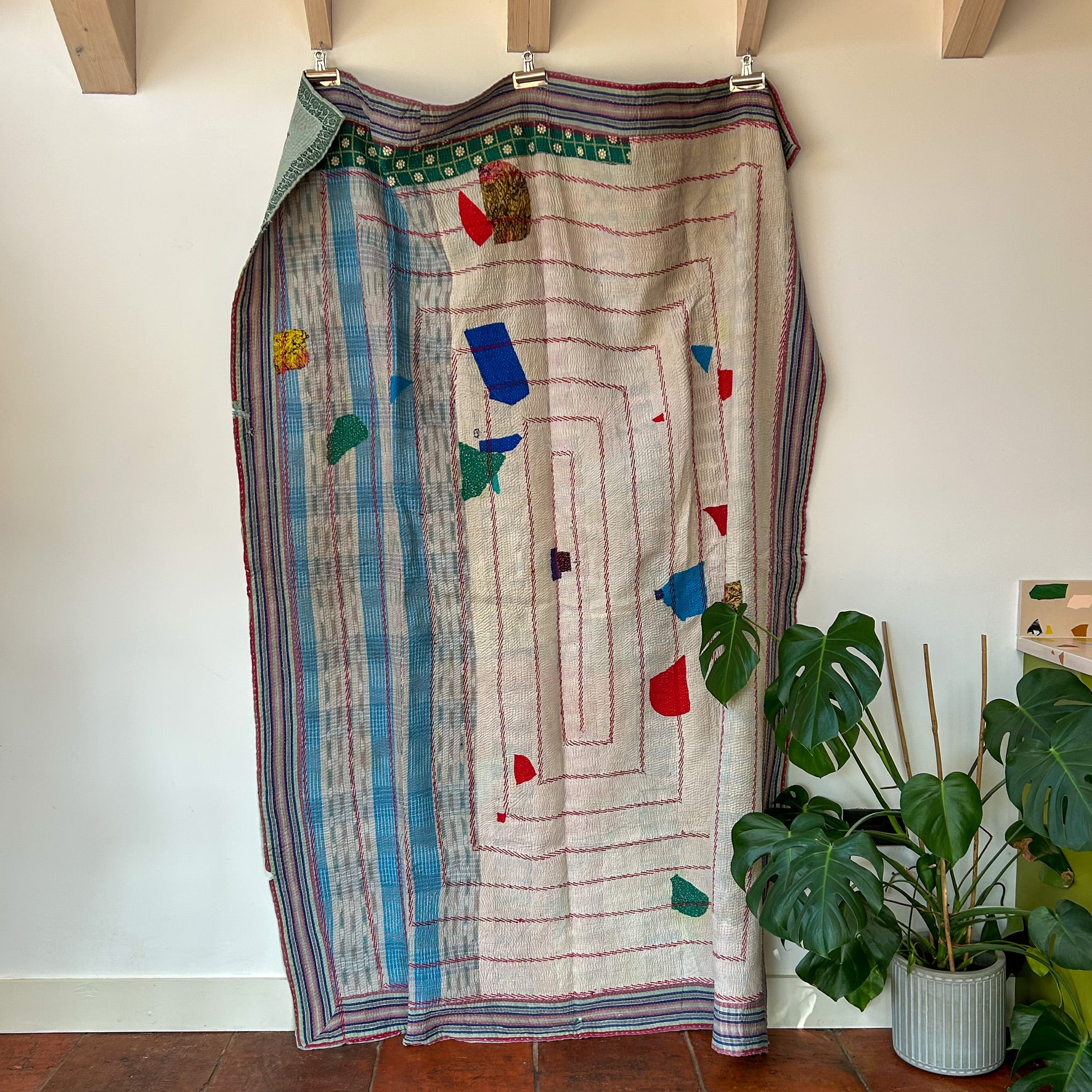 Colorful patterned skirt hanging on a rack against a white wall with a plant in the foreground. kantha-vintage-2027 - hand-made-blanket-throw-textiles-wall-hanging