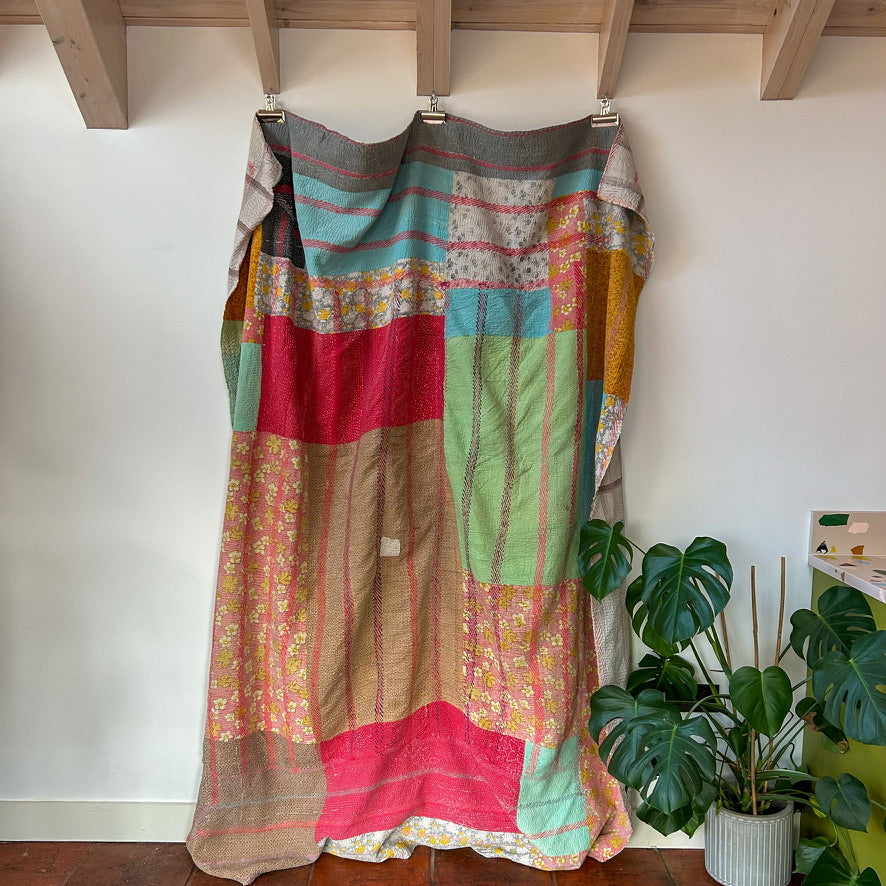 Colorful patchwork curtain hanging on a wooden rod against a white wall with plants below. kantha-vintage-2030 - hand-made-blanket-throw-textiles-wall-hanging