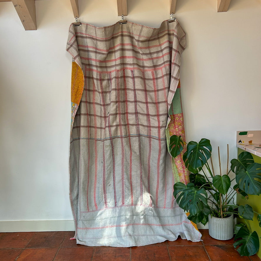 Plaid dress hanging on a rack against a white wall with plants in the background. kantha-vintage-2030 - hand-made-blanket-throw-textiles-wall-hanging