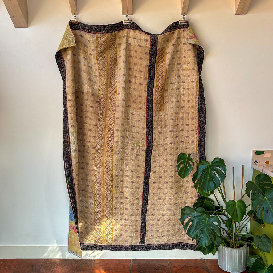 Textile with intricate patterns hanging on a rack against a white wall with a plant in the foreground. kantha-vintage-2031 - hand-made-blanket-throw-textiles-wall-hanging
