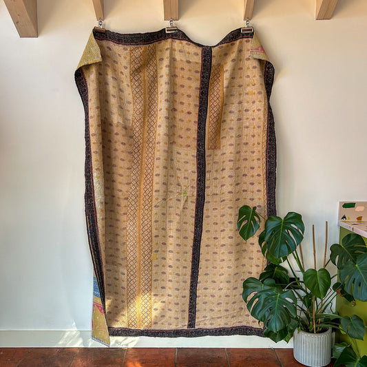 Textile with intricate patterns hanging on a rack against a white wall with a plant in the foreground. kantha-vintage-2031 - hand-made-blanket-throw-textiles-wall-hanging