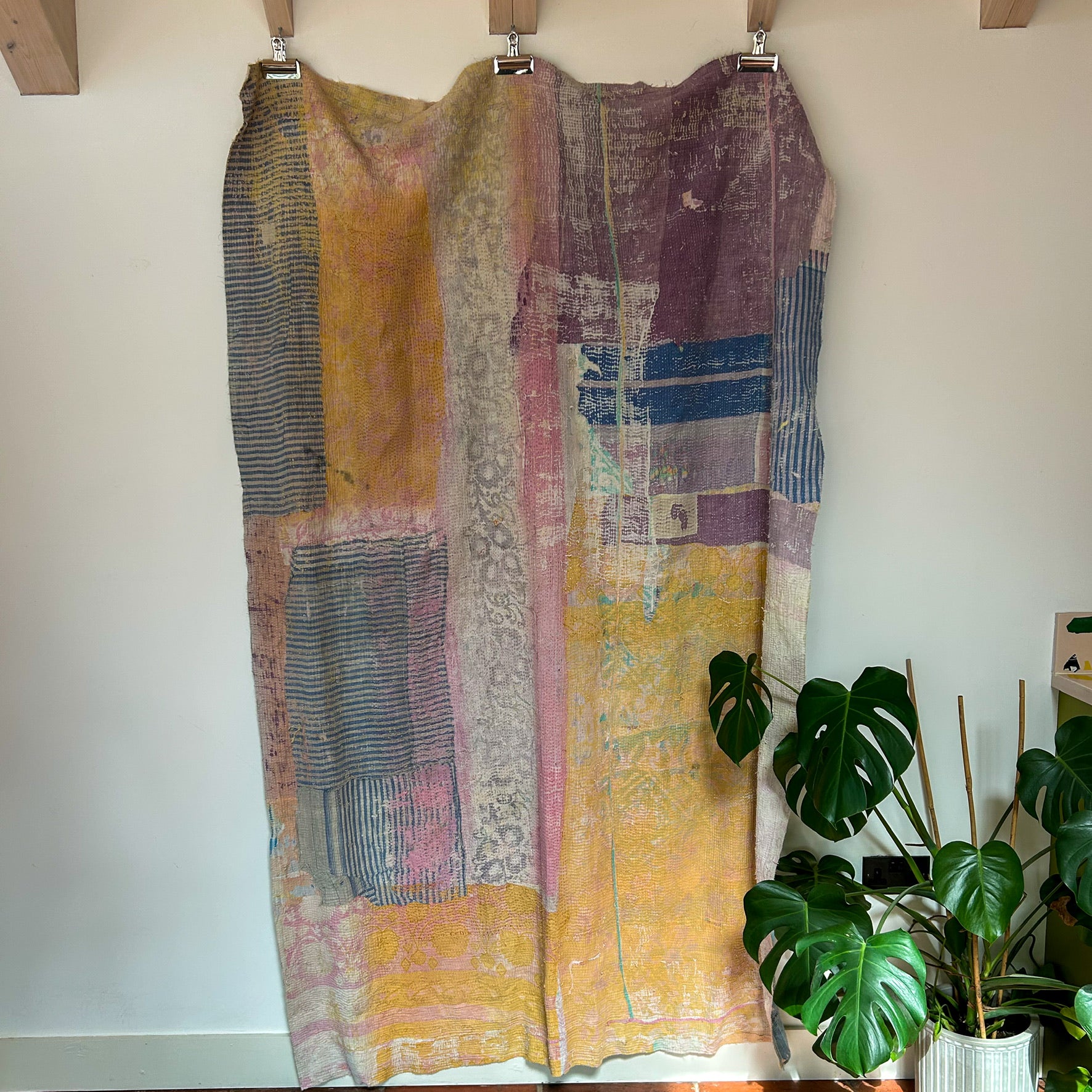 Colorful patchwork dress hanging on a rack with a plant in the background. kantha-vintage-2032 - hand-made-blanket-throw-textiles-wall-hanging