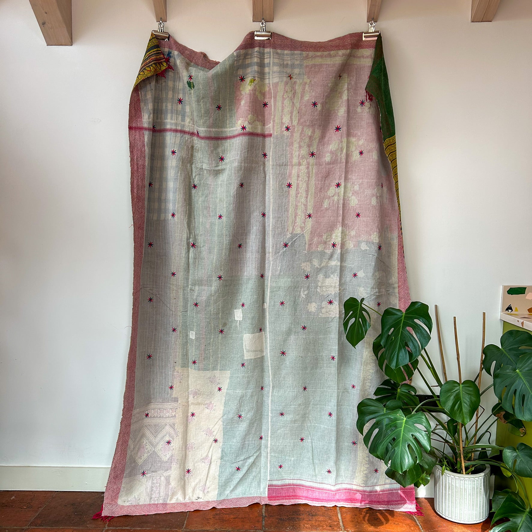 Traditional fabric draped over a wooden frame against a white wall with a plant in the foreground.  kantha-vintage-2035- hand-made-blanket-throw-textiles-wall-hanging