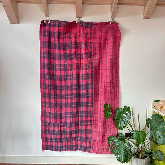 Red and black plaid curtain hanging on a wooden rod against a white wall with a plant in the foreground.  kantha-vintage-2036- hand-made-blanket-throw-textiles-wall-hanging