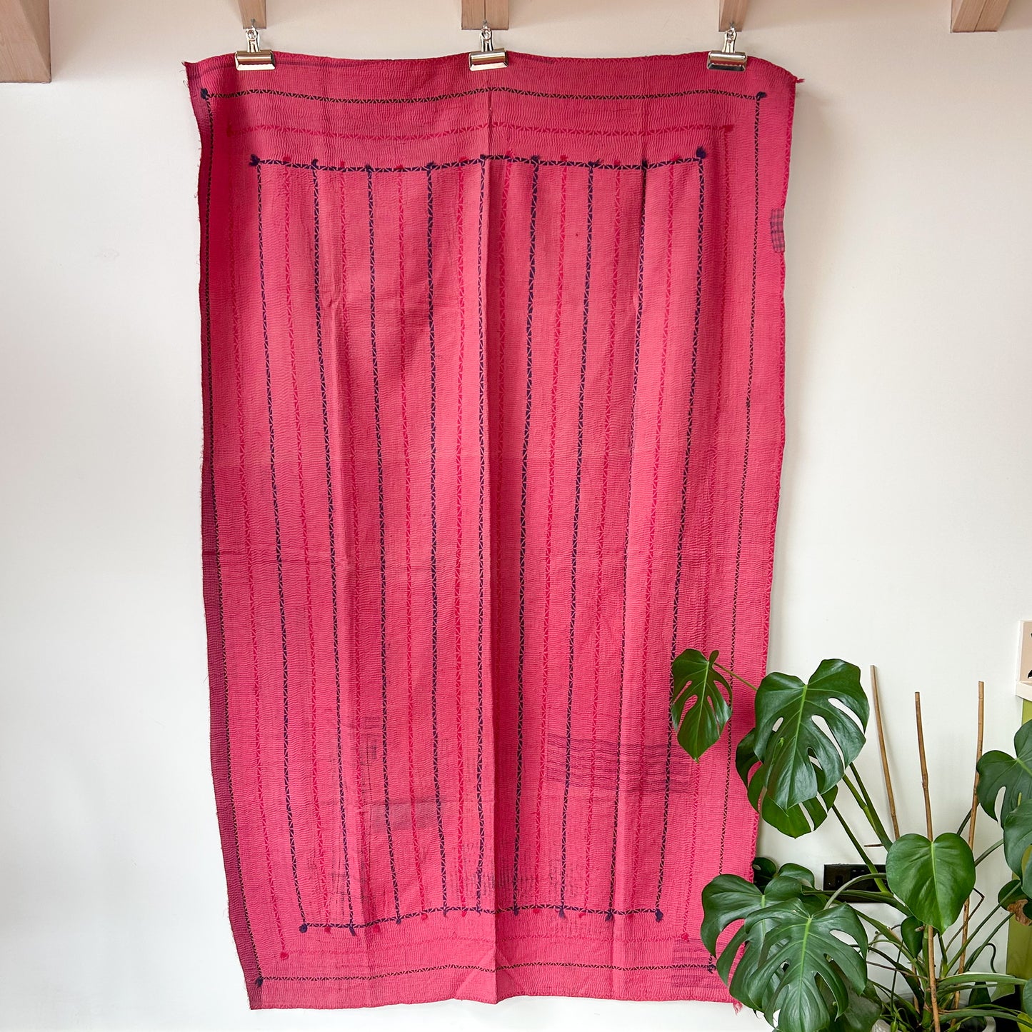 Pink curtain with black stripes hanging on a wooden rod against a white wall with plants in the background.  kantha-vintage-2036- hand-made-blanket-throw-textiles-wall-hanging