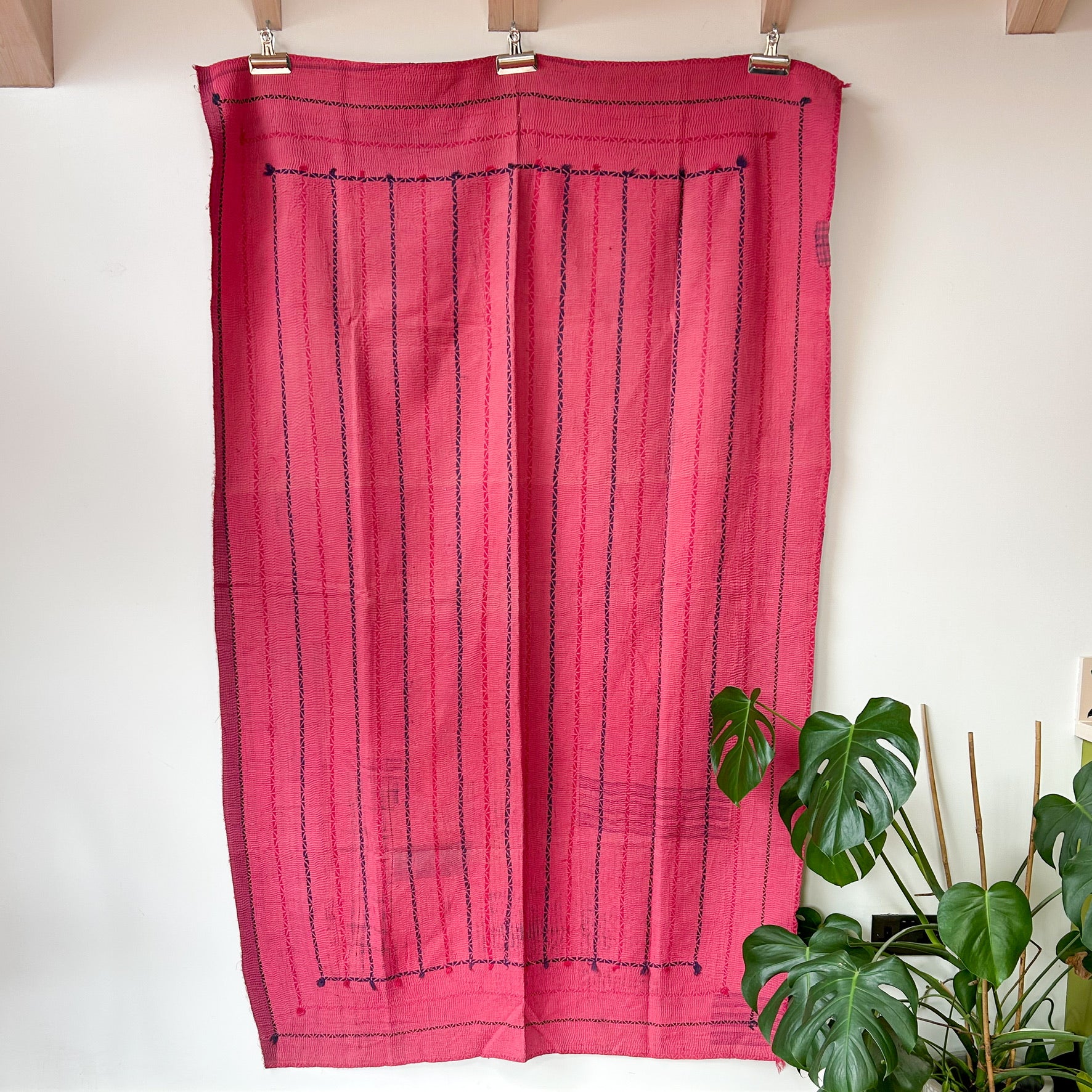 Pink curtain with black stripes hanging on a wooden rod against a white wall with plants in the background.  kantha-vintage-2036- hand-made-blanket-throw-textiles-wall-hanging