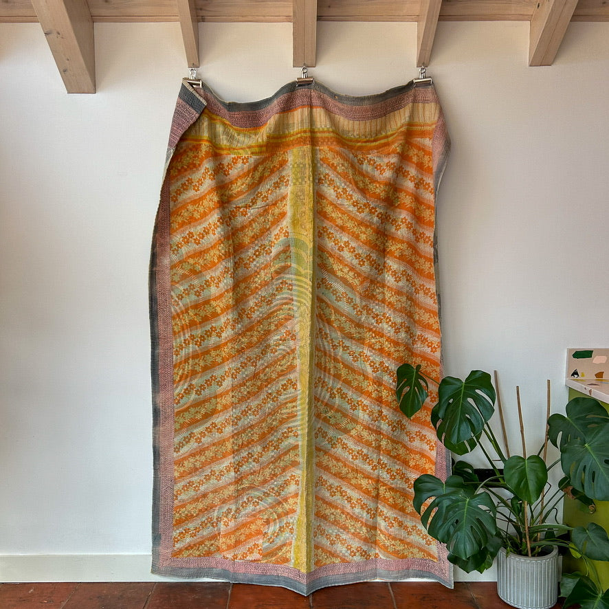 Colorful patterned fabric hanging on a wooden rod against a white wall with a plant in the corner. 