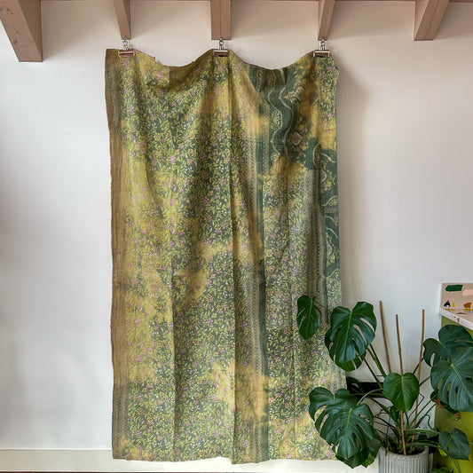 Green patterned curtain hanging on a wooden rod against a white wall with a plant in the foreground.