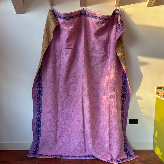 Pink fabric kantha with purple border on a wooden hanger against a white wall.