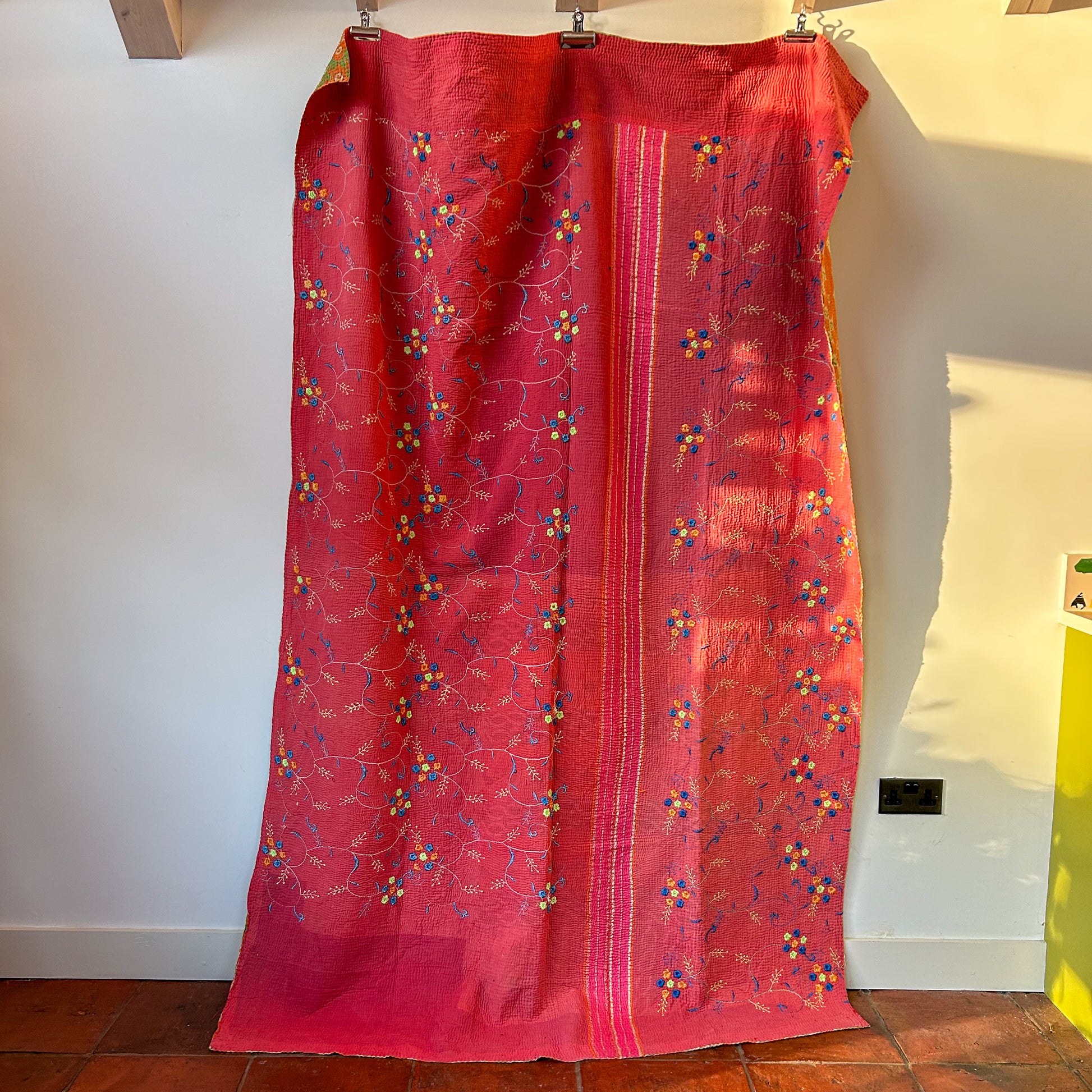 Red kantha with floral patterns hanging on a wooden rod against a white wall.