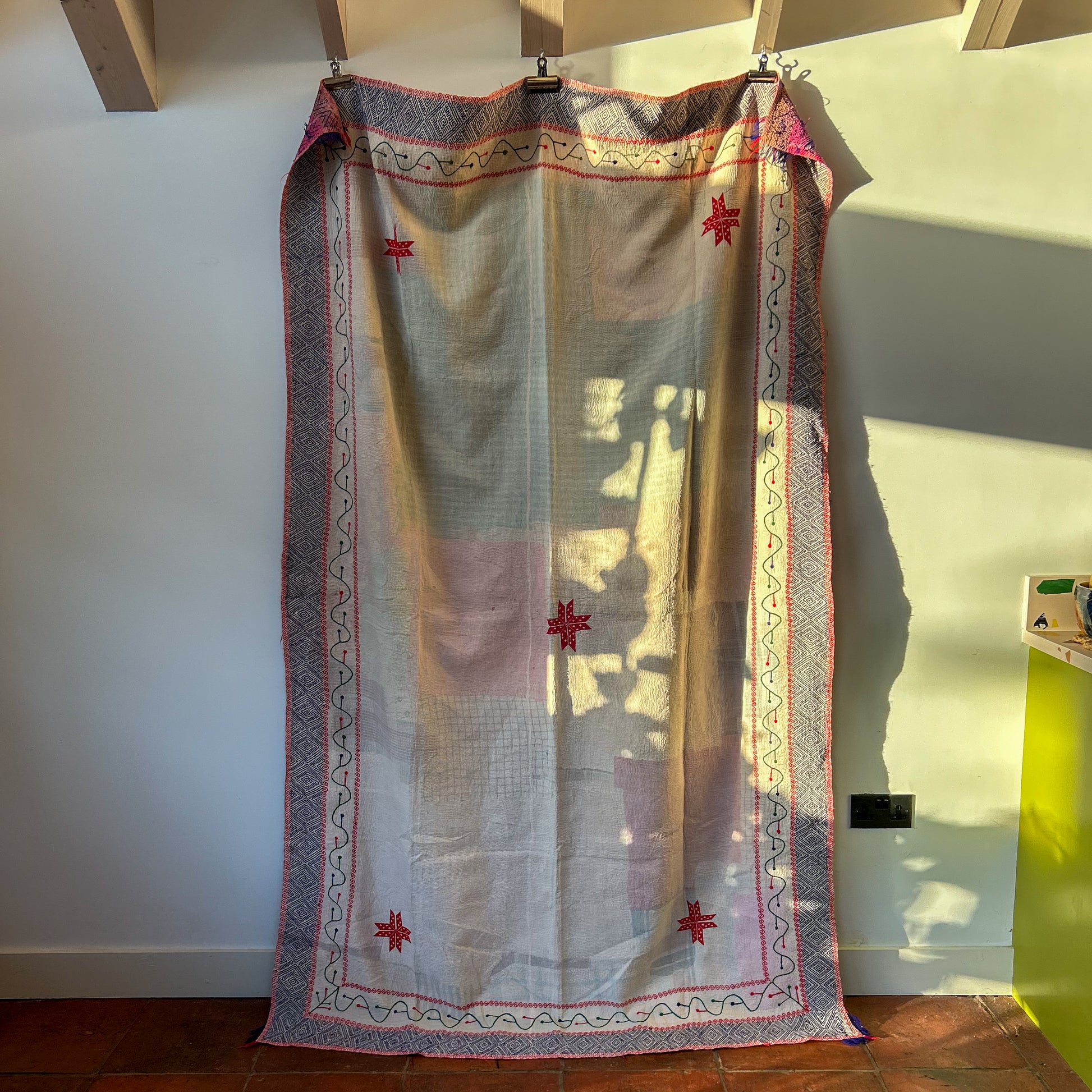 Embroidered kantha with red star patterns hanging on a wooden rod against a white wall.