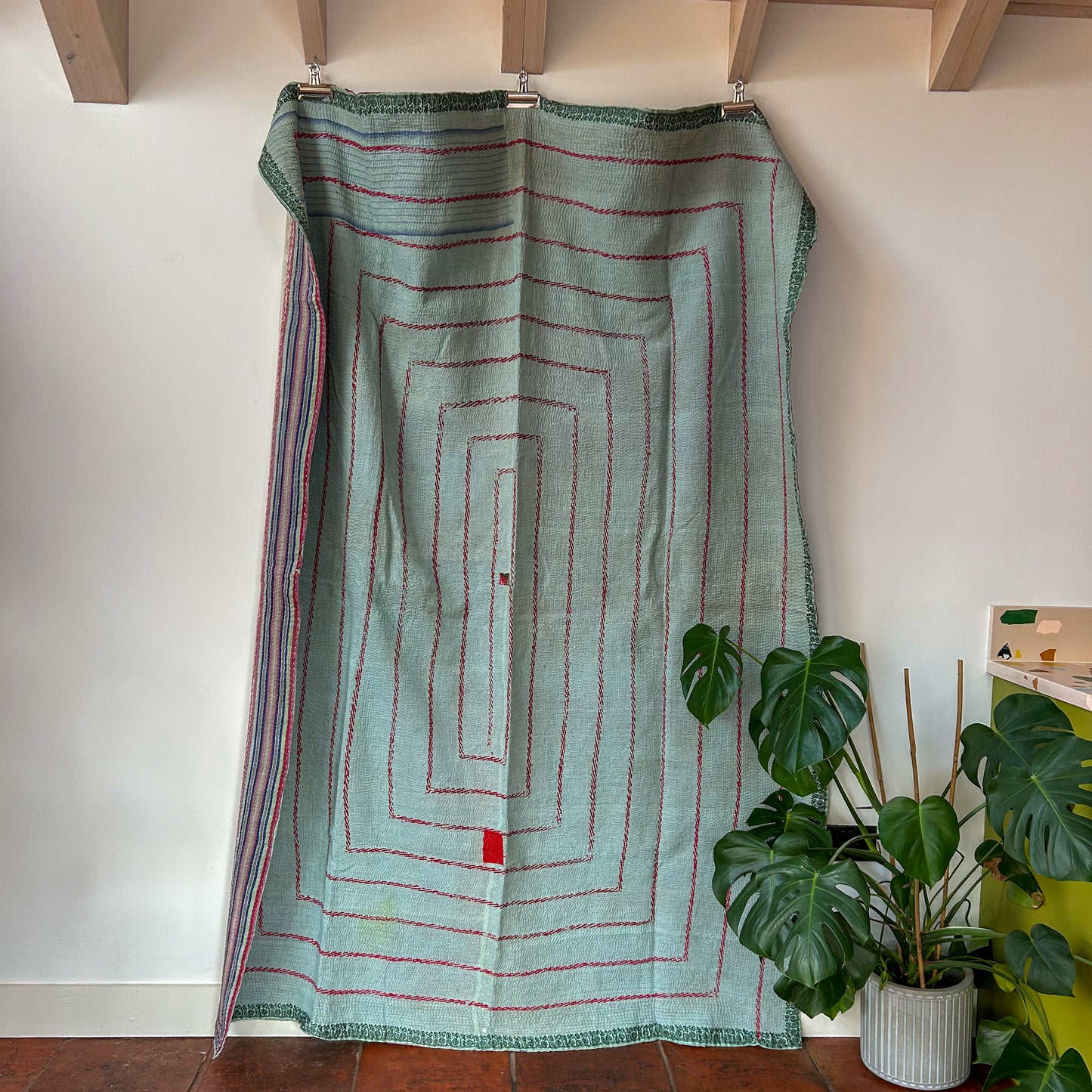 Textile with red patterns draped over a wooden frame against a white wall with plants below. kantha-vintage-2027 - hand-made-blanket-throw-textiles-wall-hanging