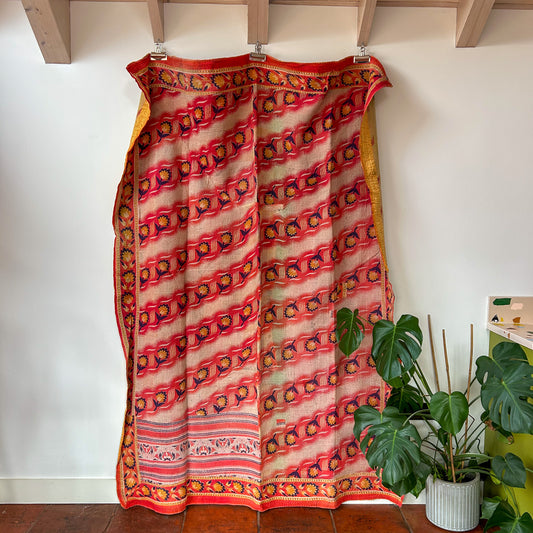 Traditional patterned fabric draped over a wooden frame against a white wall with a plant in the corner. kantha-vintage-2011-hand-made-blanket-throw-textiles-wall-hanging