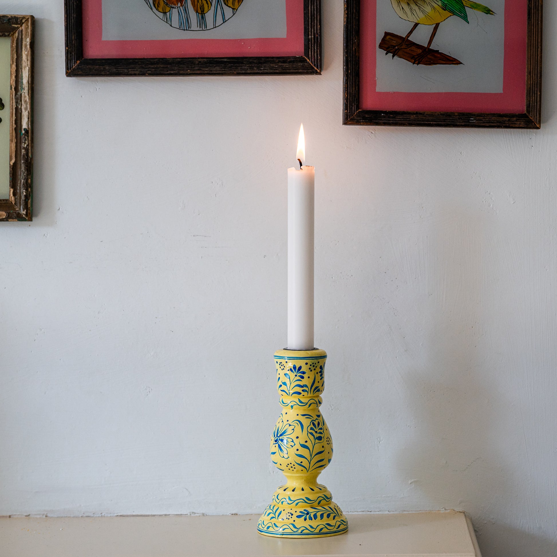 Yellow candle holder with a white candle on a white surface, framed pictures in the background. constance-and-denny-candle-stick-holder-cream-hand-painted-wooden