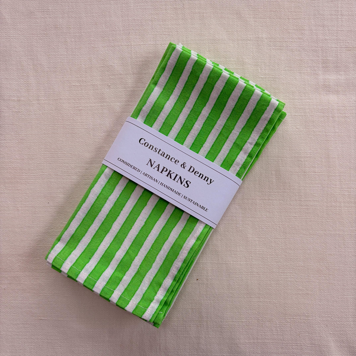 A folded set of Constance & Denny napkins with bright green and white vertical stripes, wrapped in a white paper band label, displayed on a textured beige linen background.