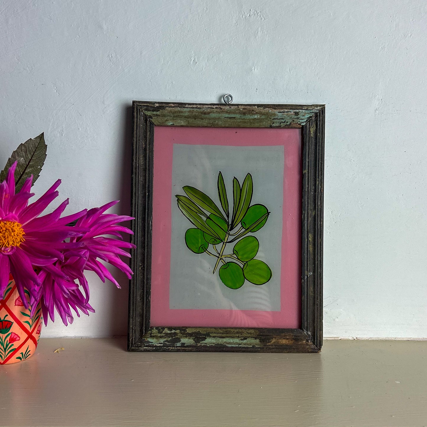 Framed artwork with green leaves on a pink background next to a purple flower in a colorful vase.
