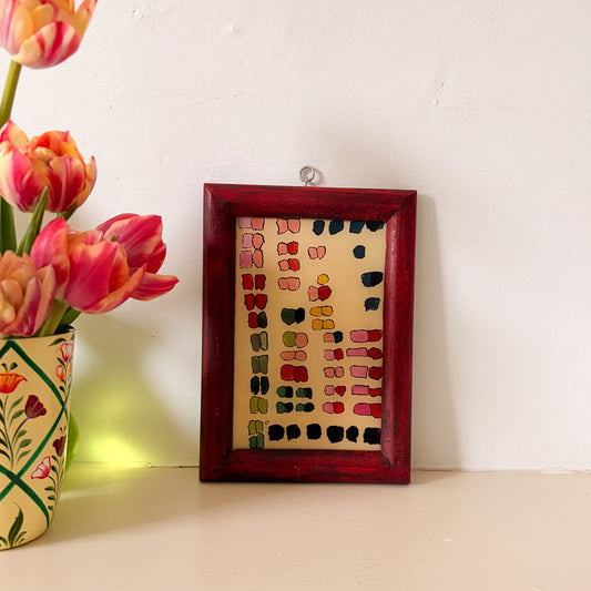 Small framed artwork with colorful patterns next to a vase of pink tulips on a white surface.