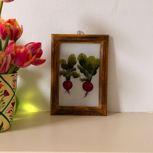Framed artwork of two radishes on a white surface next to a vase of pink tulips.