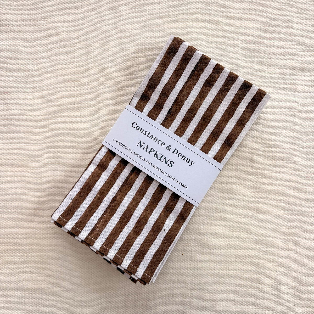 A stack of brown and white striped paper napkins wrapped with a white paper band label that reads 'Constance & Denny NAPKINS', placed on a textured cream linen background.