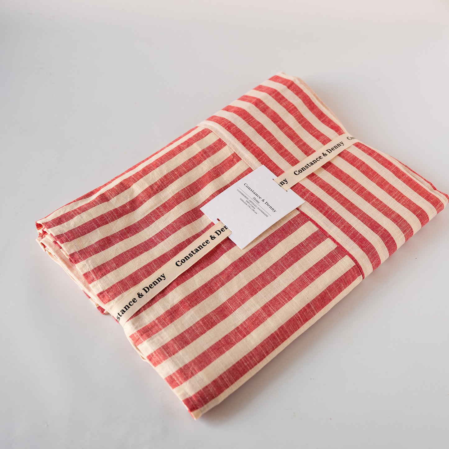 A folded striped fabric item, likely a tablecloth or napkin set, featuring alternating red and cream horizontal stripes. It is wrapped with a paper band labeled 'Constant & Denny' and a small white tag, photographed on a clean white background.