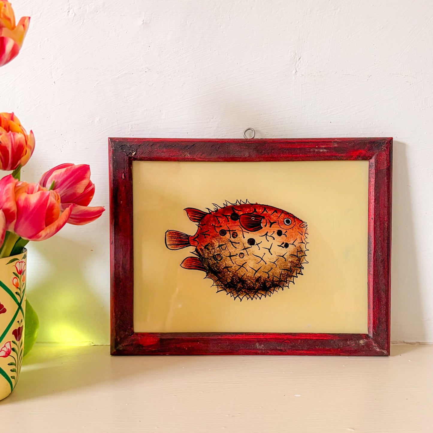 Framed artwork of a puffer fish on a beige background with flowers to the left. red puffer fish indian glass paintng