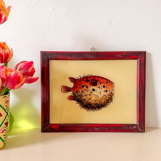 Framed artwork of a puffer fish on a beige background with flowers to the left. red puffer fish indian glass paintng