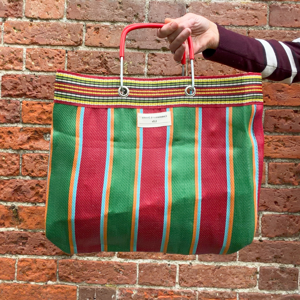 A striped woven tote bag with red, green, orange, and blue vertical stripes, held by a person's hand against a red brick wall background.