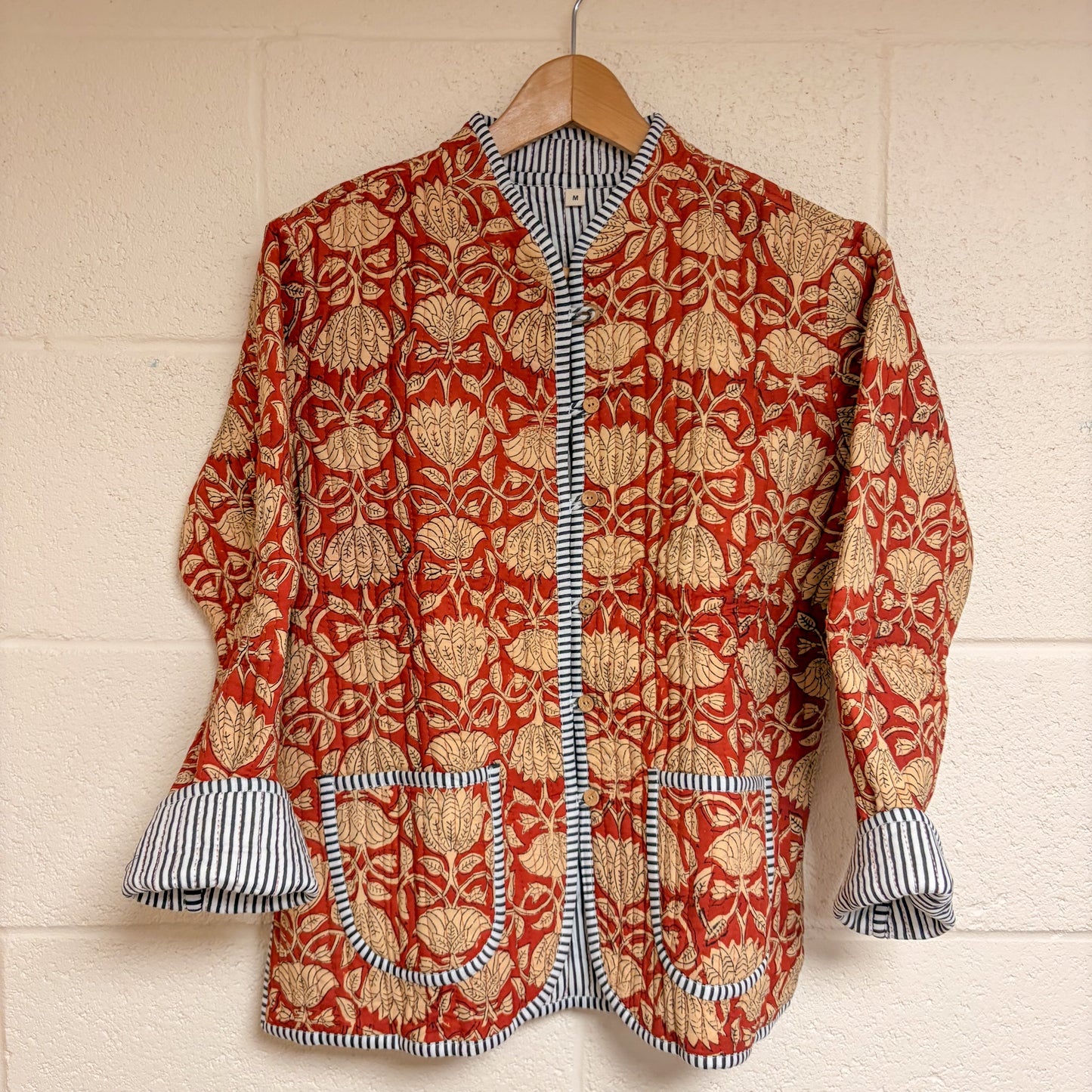 Red and gold patterned jacket with striped cuffs and trim on a wooden hanger against a beige wall.