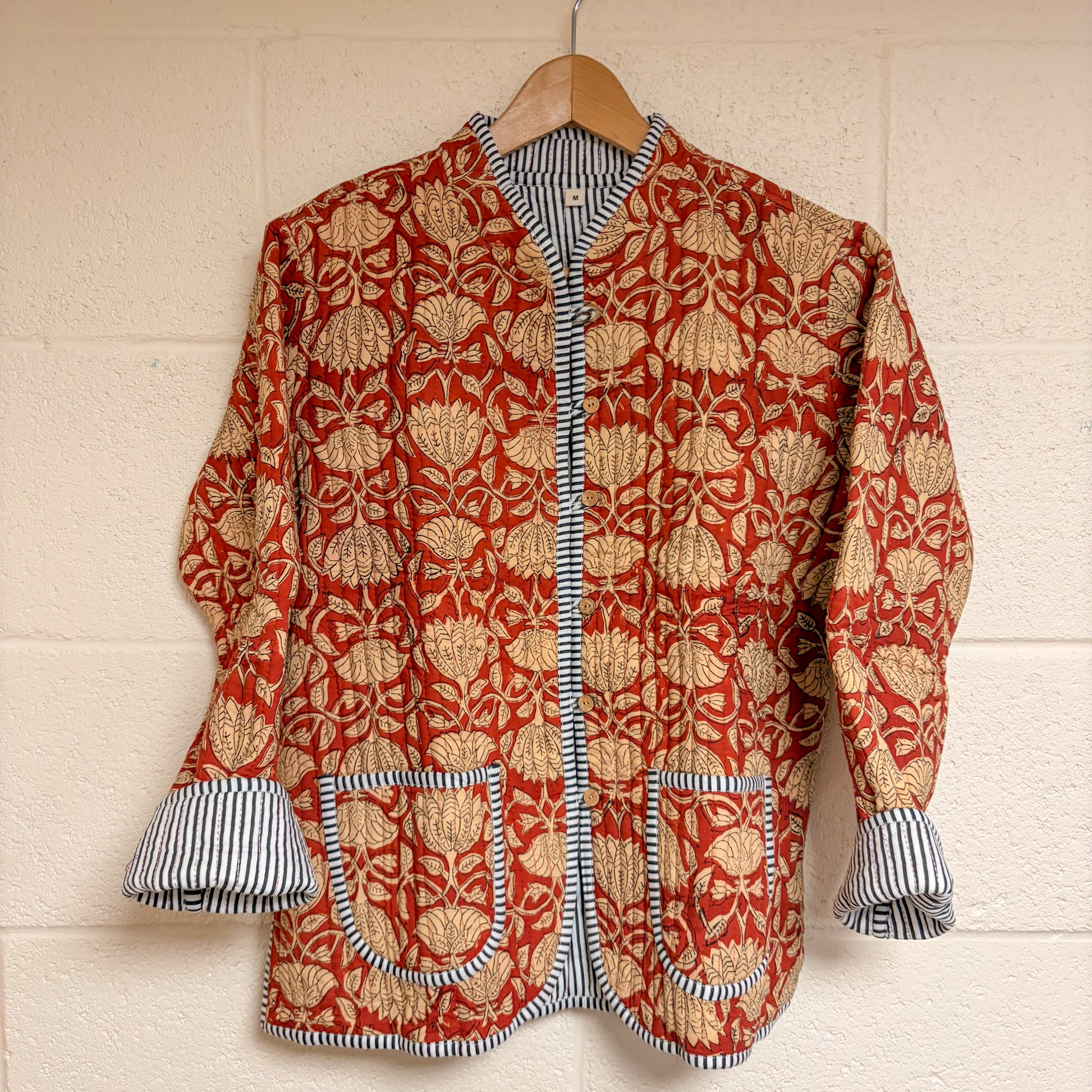 Red and gold patterned jacket with striped cuffs and trim on a wooden hanger against a beige wall.