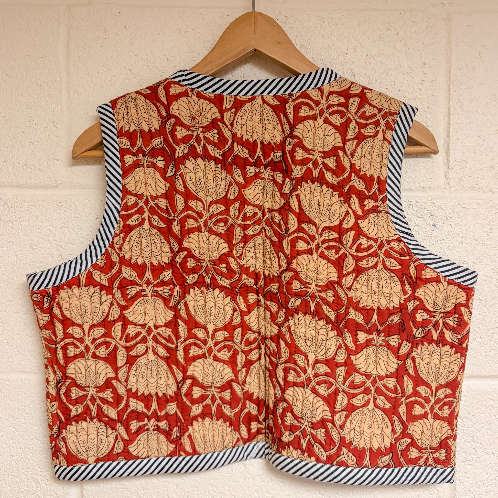 Red floral patterned vest with black and white striped trim on a wooden hanger against a beige wall.